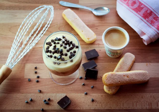 some of the ingredients for this tiramisu without alcohol recipe: savoiardi biscuits, coffee and chocolate, next to a whisk.