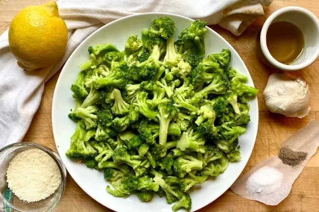 the ingredients for this sauteed frozen broccoli recipe on a table: broccoli, a lemon, garlic cloves, parmesan, and spices