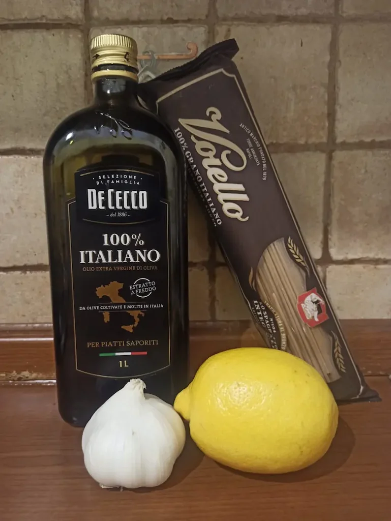 all the ingredients for this lemon pasta recipe: a lemon, garlic cloves, spaghetti, and extra virgin olive oil