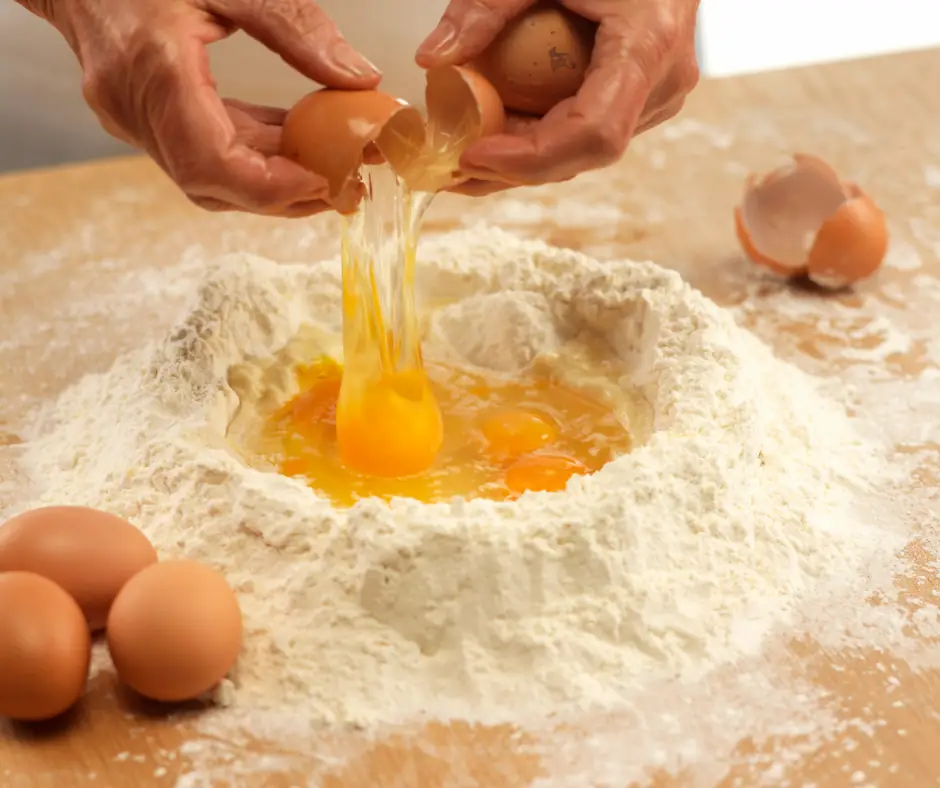 detail of two hands breaking eggs onto a mountain of flour on a large work surface