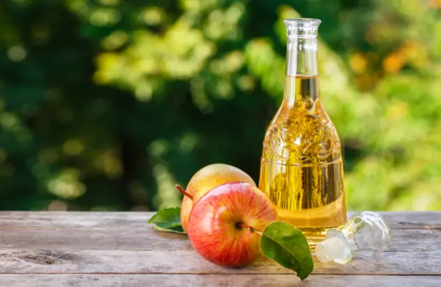 a bottle of apple cider on a table in the open air, aside two apples