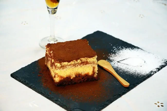 a finished piece of tiramisu without alcohol on a black plate, with sprinkels of cocoa powder and sugar