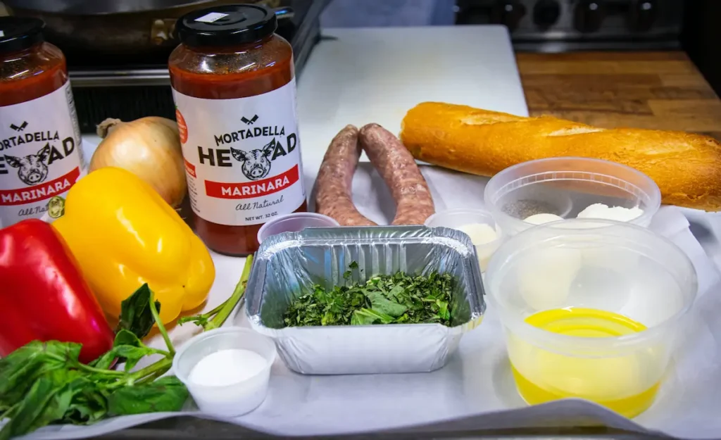the ingredients for this baked sausage recipe on a work surface: bell peppers, fresh greens, Mortadella Head's marinara sauce, salt, oil, and of course Italian sausages