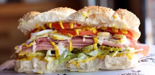 a very big sandwich with cold cut meat, greens and tomatoes