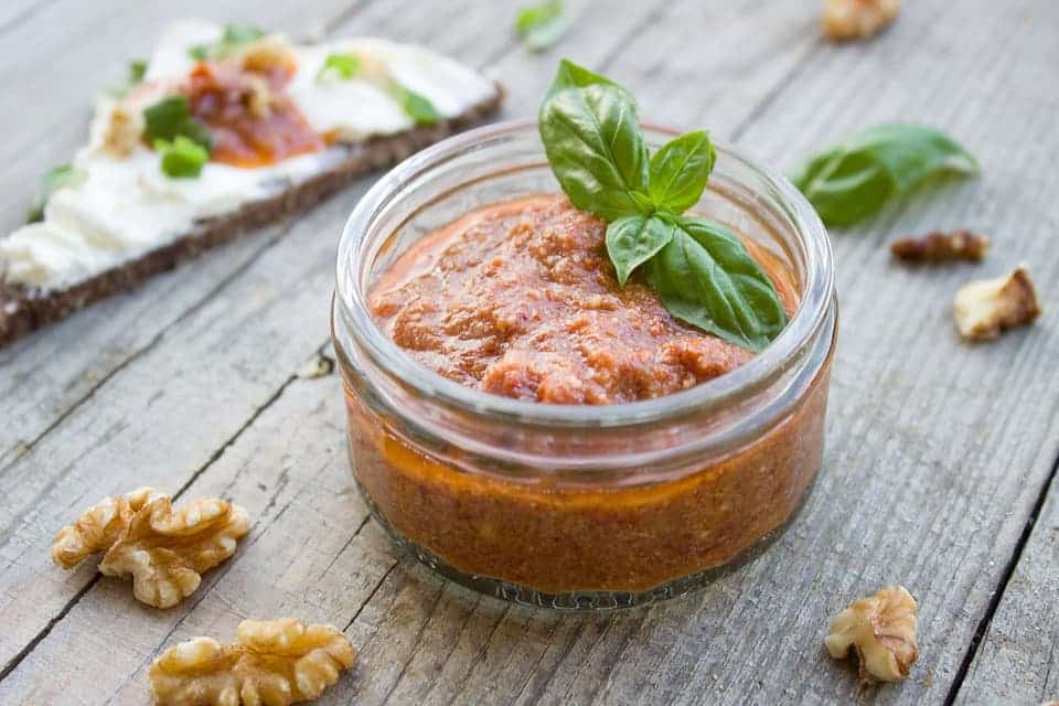 a little jar full of tomato pesto garnished with a bunch of basil leaves, left open on a table with some walnuts and a bruschetta in the background