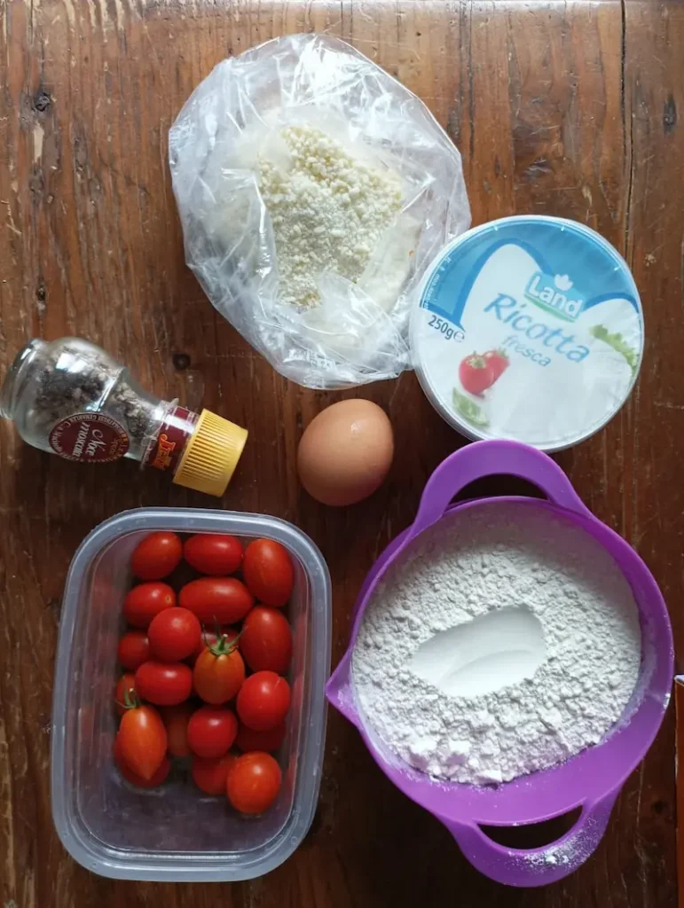 the ingredients for this ricotta gnocchi recipe on a table: ricotta, flour, an egg, parmesan cheese, nutmeg, and some date tomatoes for the dressing