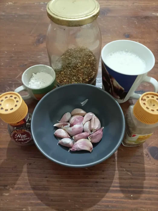 some of the ingredients for this garlic pizza sauce recipe on a wooden table: garlic cloves, nutmeg, black pepper, flour, milk, and Italian seasoning