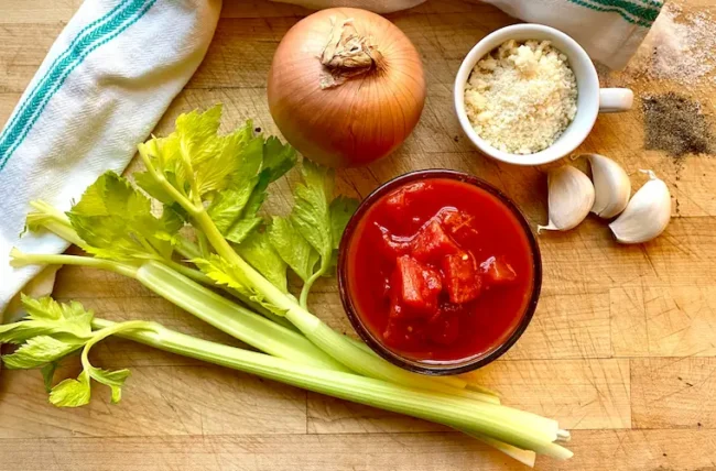 the ingredients for this recipe on a table: celery sticks, an onion, peeled tomatoes, garlic and parmesan cheese